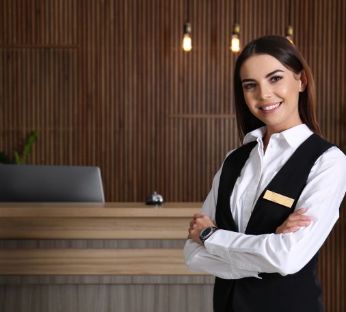 "Person in white shirt and black vest with name tag, standing with arms crossed in front of a reception desk with wooden paneling and hanging lights.