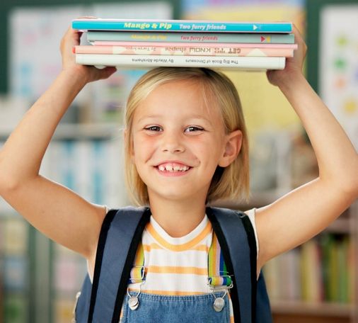 Smiling young girl with a backpack balancing a stack of colorful books on her head in a cheerful classroom setting.