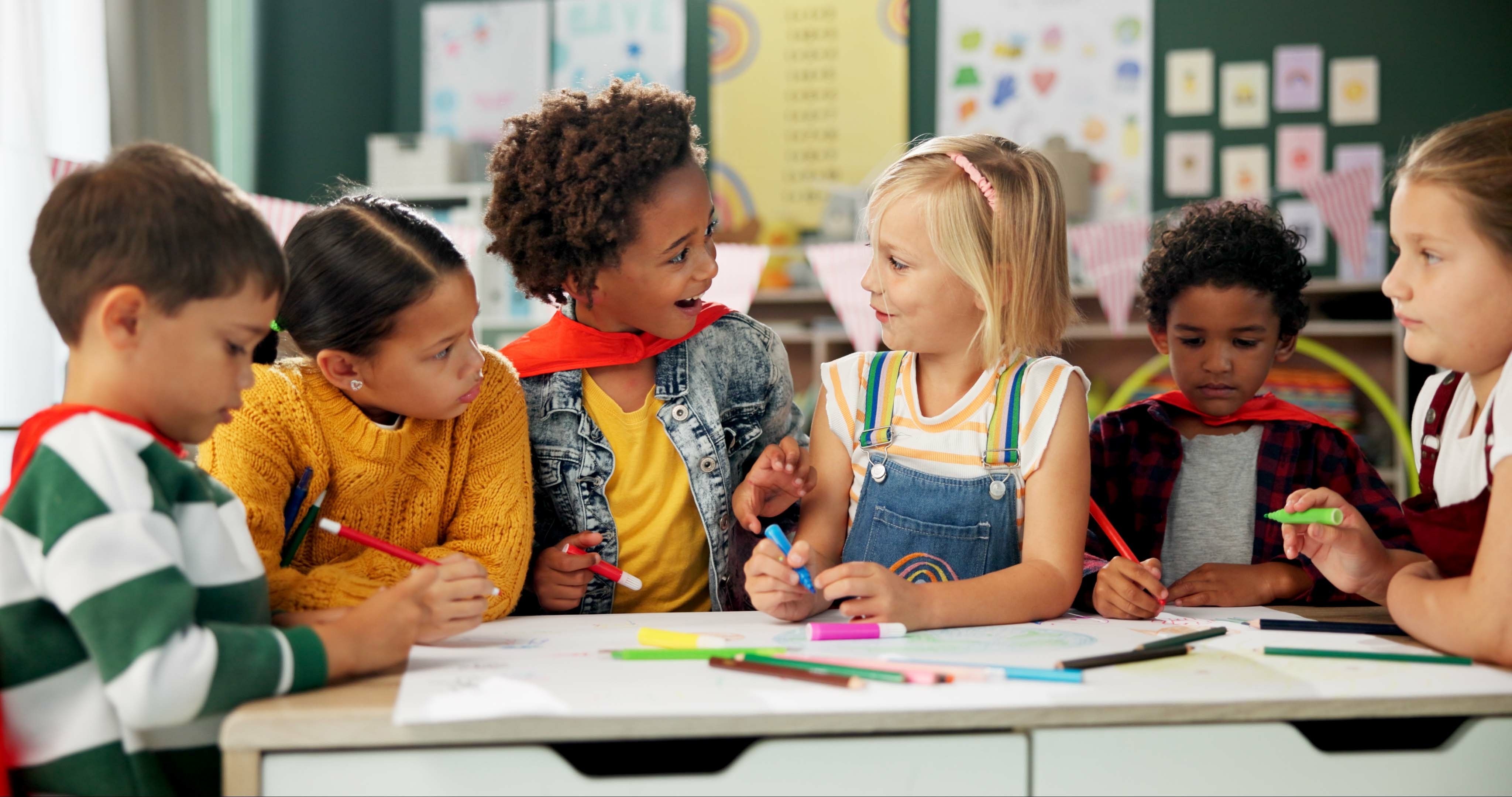 Group of young children in a colorful classroom engaged in a creative activity, drawing and chatting together at a table.