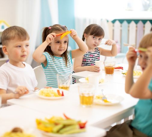 Young children sitting at a table in a bright and colorful cafeteria, eating healthy meals with fresh vegetables and juice, while some playfully interact with their food.