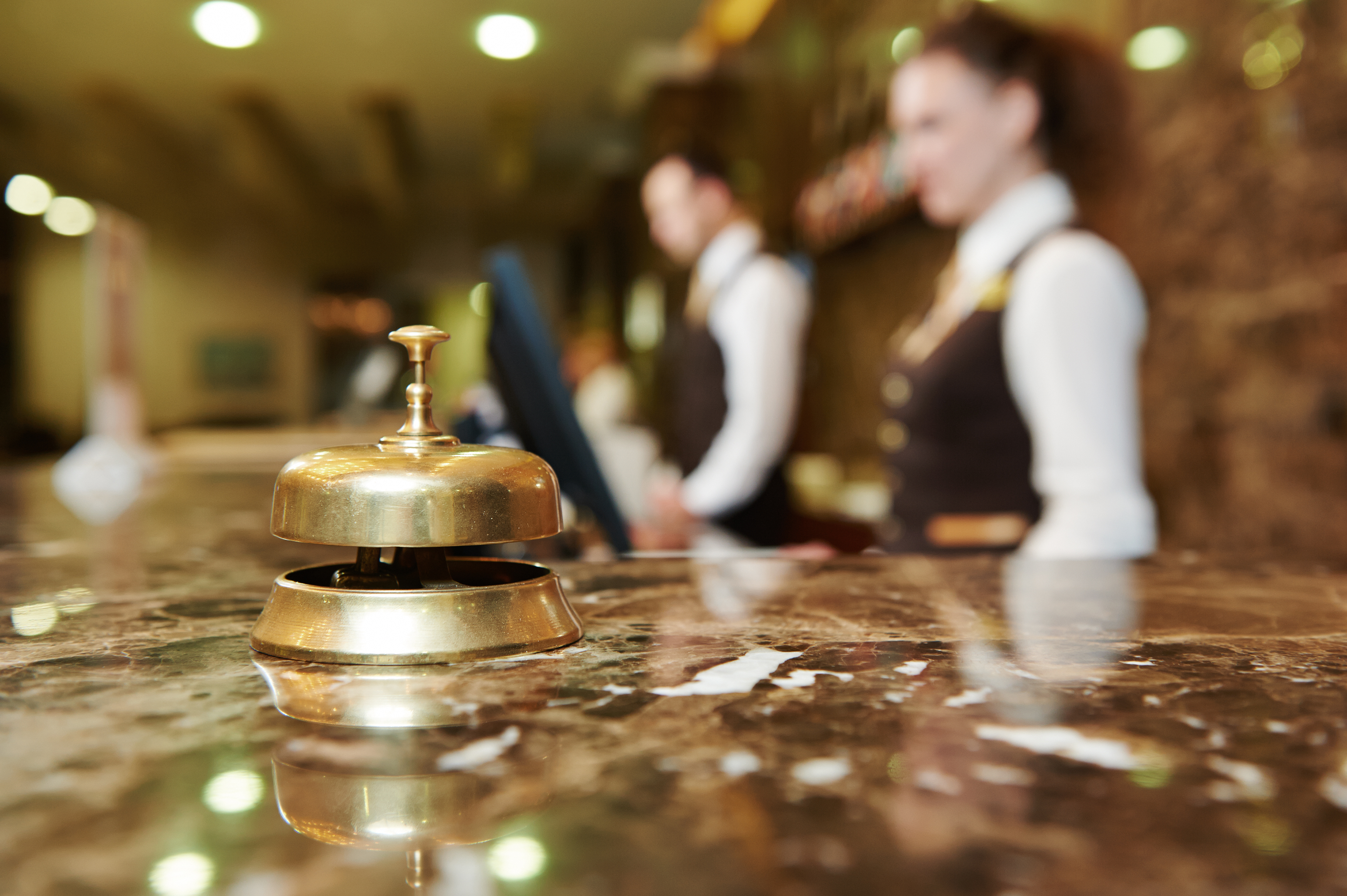 Hotel reception desk with brass service bell in focus. Two staff members in uniform behind the counter, one using a computer.